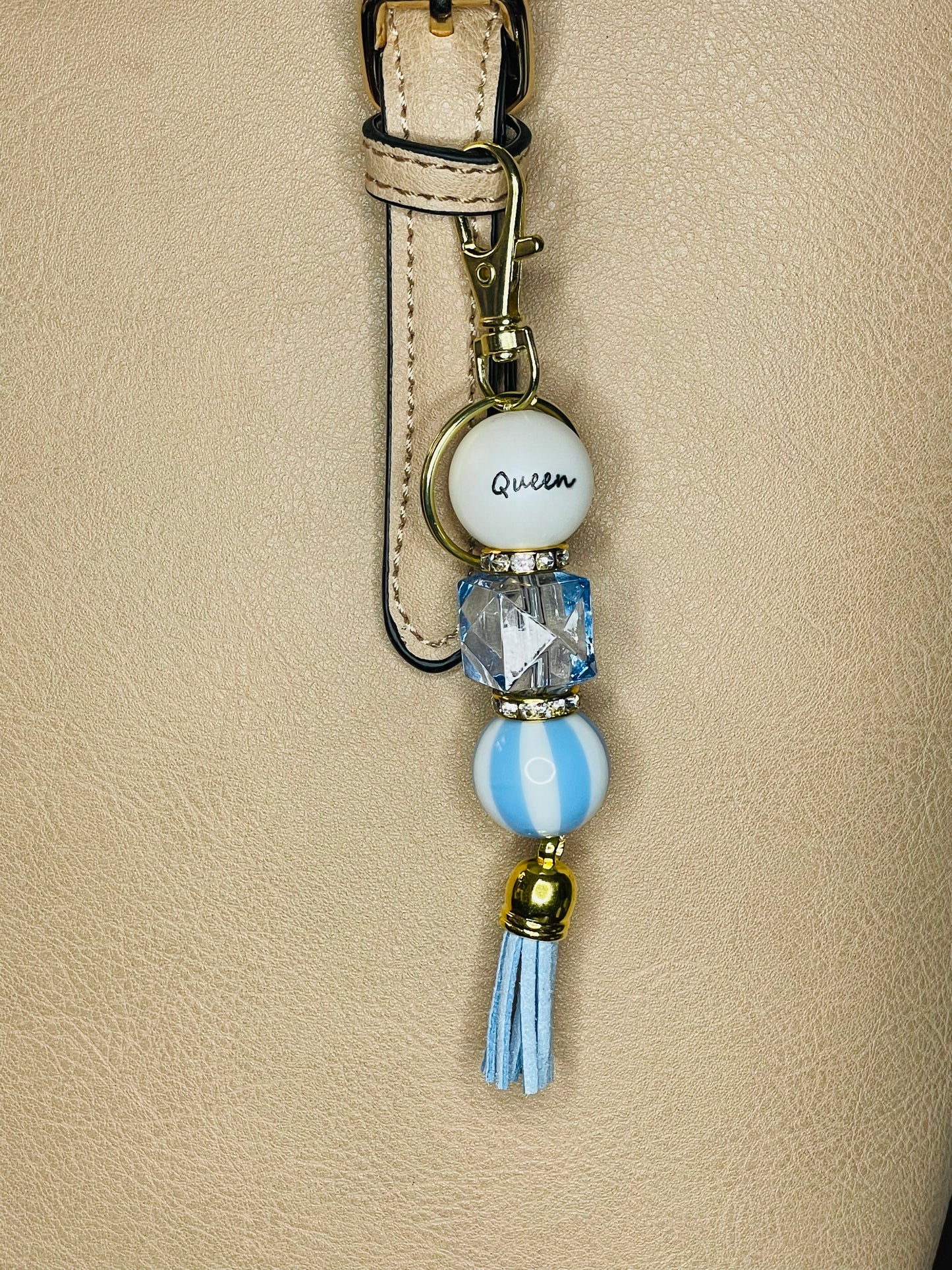 A key ring on a gold ring with 3 with gold rhinestone spacers between each, beads the first white with Queen written on the bead, the second is a clear light blue square bead, the third is light blue and white vertical striped. The key chain is finished with a light blue tassel..