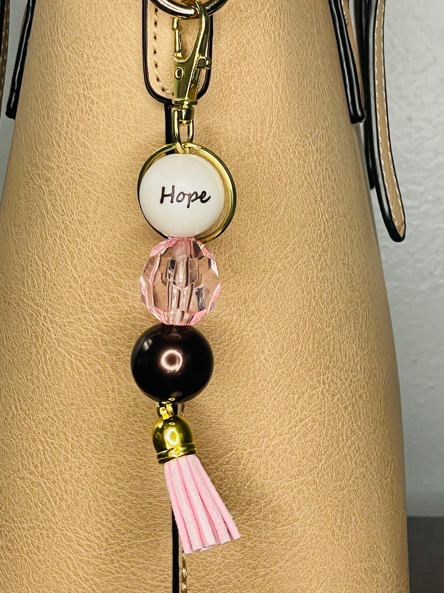A key ring on a gold ring with 3 beads the first white with hope written on the bead, the second is clear pink facetted, the third a deep chocolate shiny brown. The key chain is finished with a pink tassel.