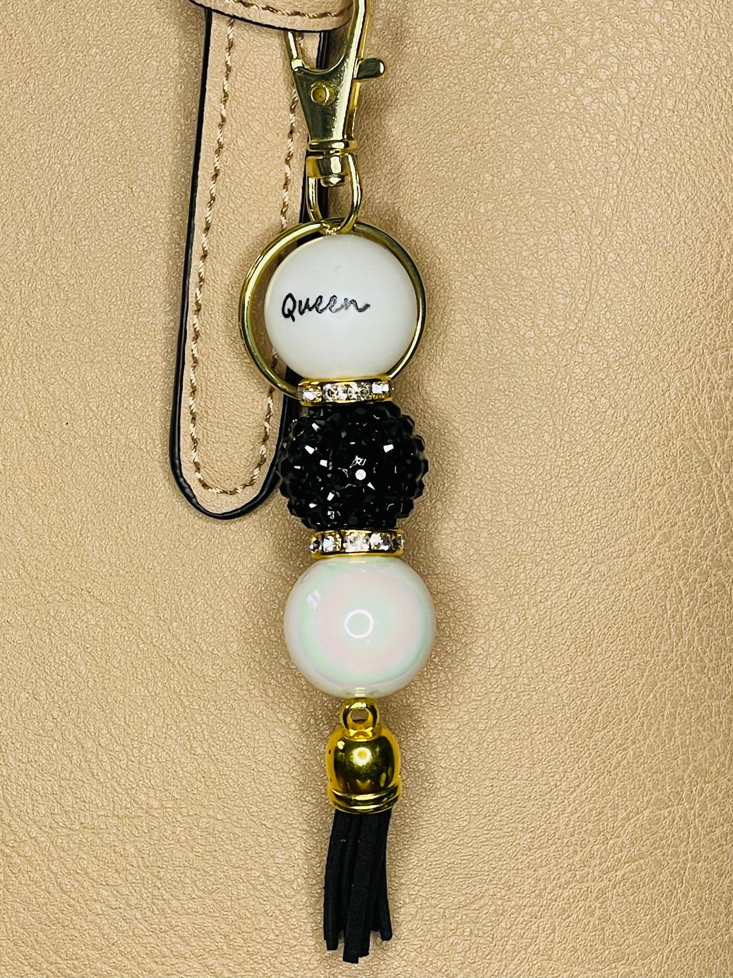 A key ring on a gold ring with 3 with gold rhinestone spacers between each, beads the first white with Queen written on the bead, the second is a black rhinestone bead, the third is an iridescent pearl bead. The key chain is finished with a black tassel.