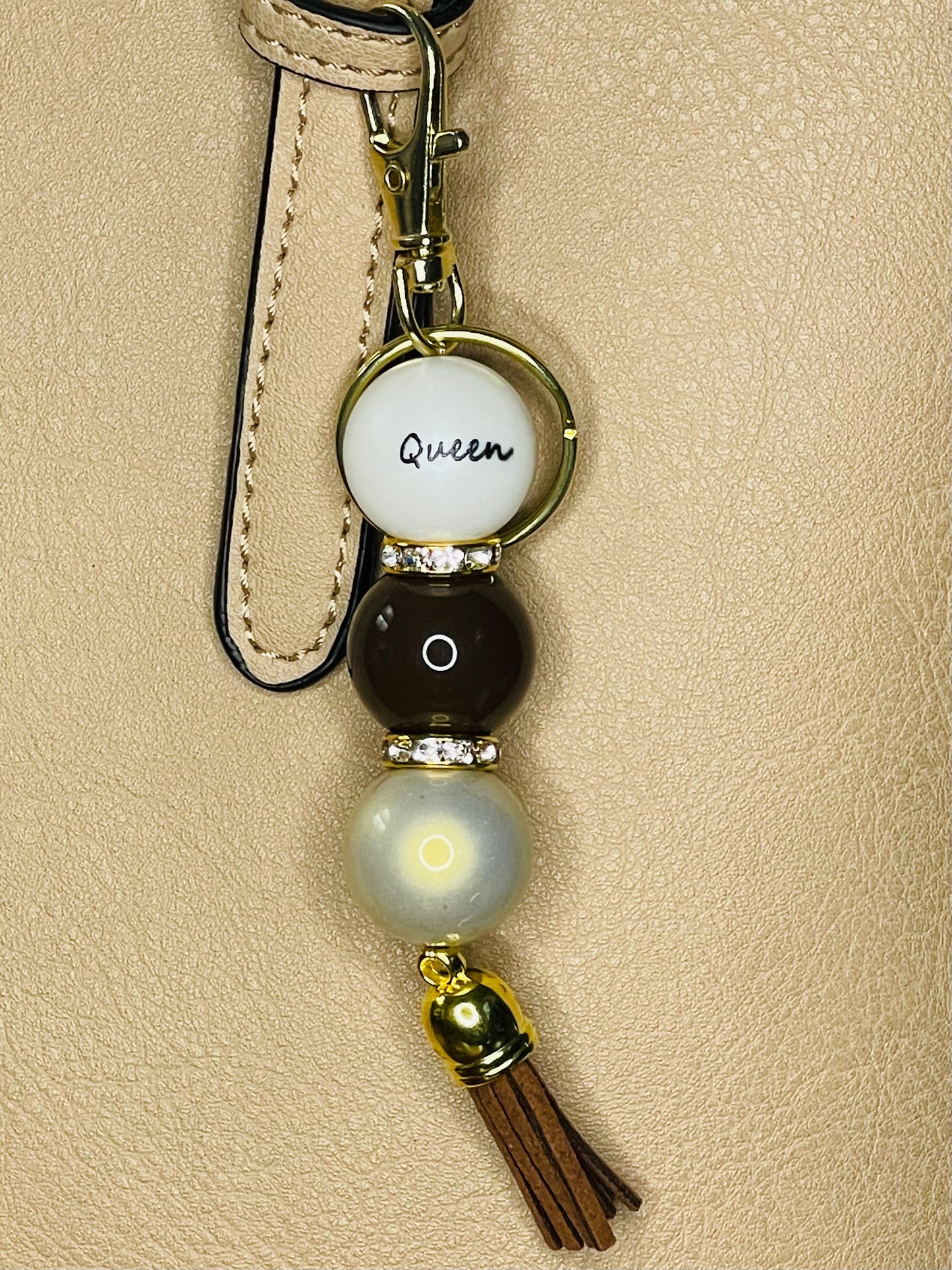 A key ring on a gold ring with 3 with gold rhinestone spacers between each, beads the first white with Queen written on the bead, is a chocolate, the third a shiny light gold . The key chain is finished with a brown tassel.