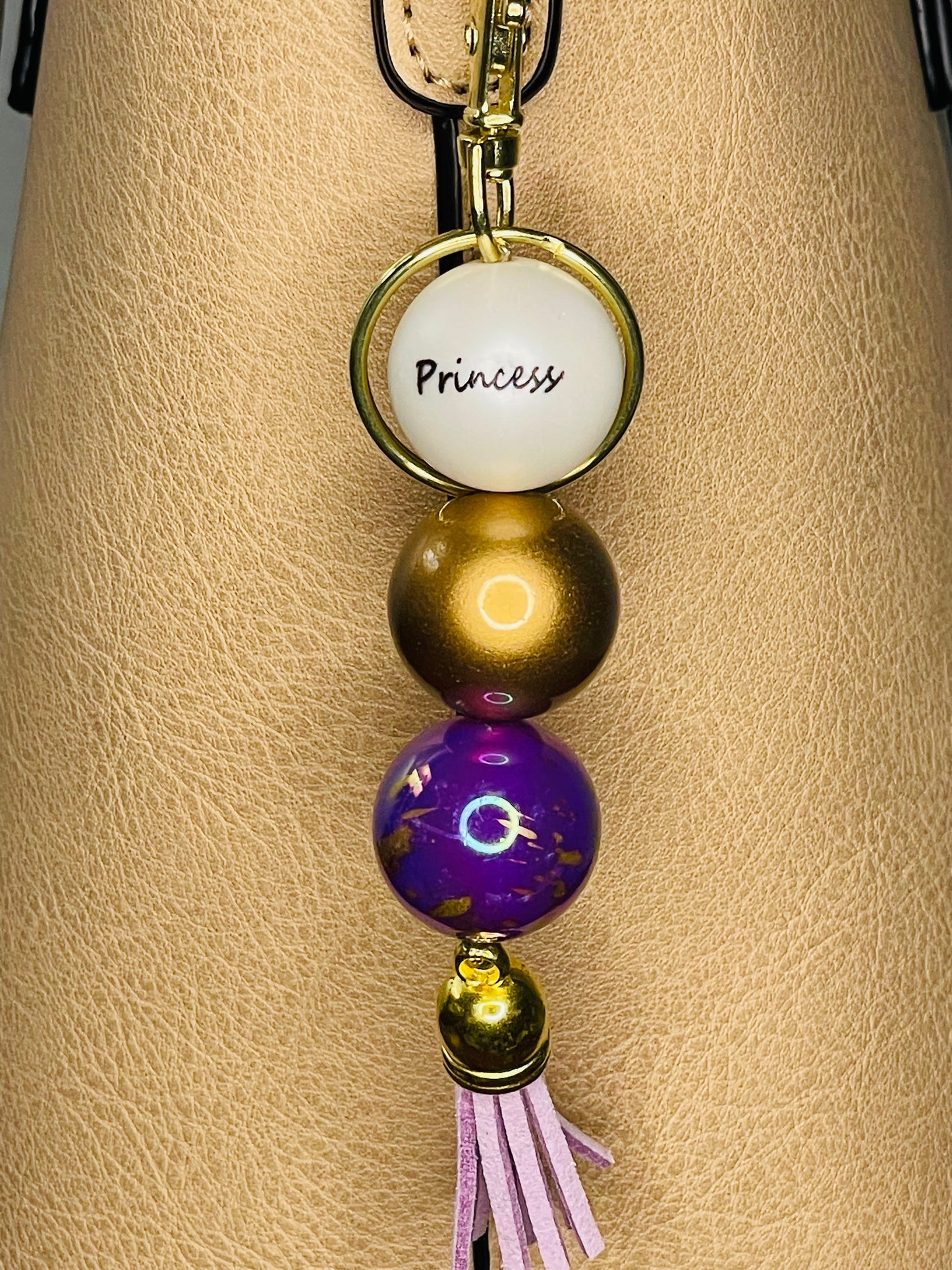 A key ring on a gold ring with 3 beads the first white with Princess written on the bead, is a deep shiny gold , the third a dark purple with gold fleck swirls. The key chain is finished with a light purple tassel
