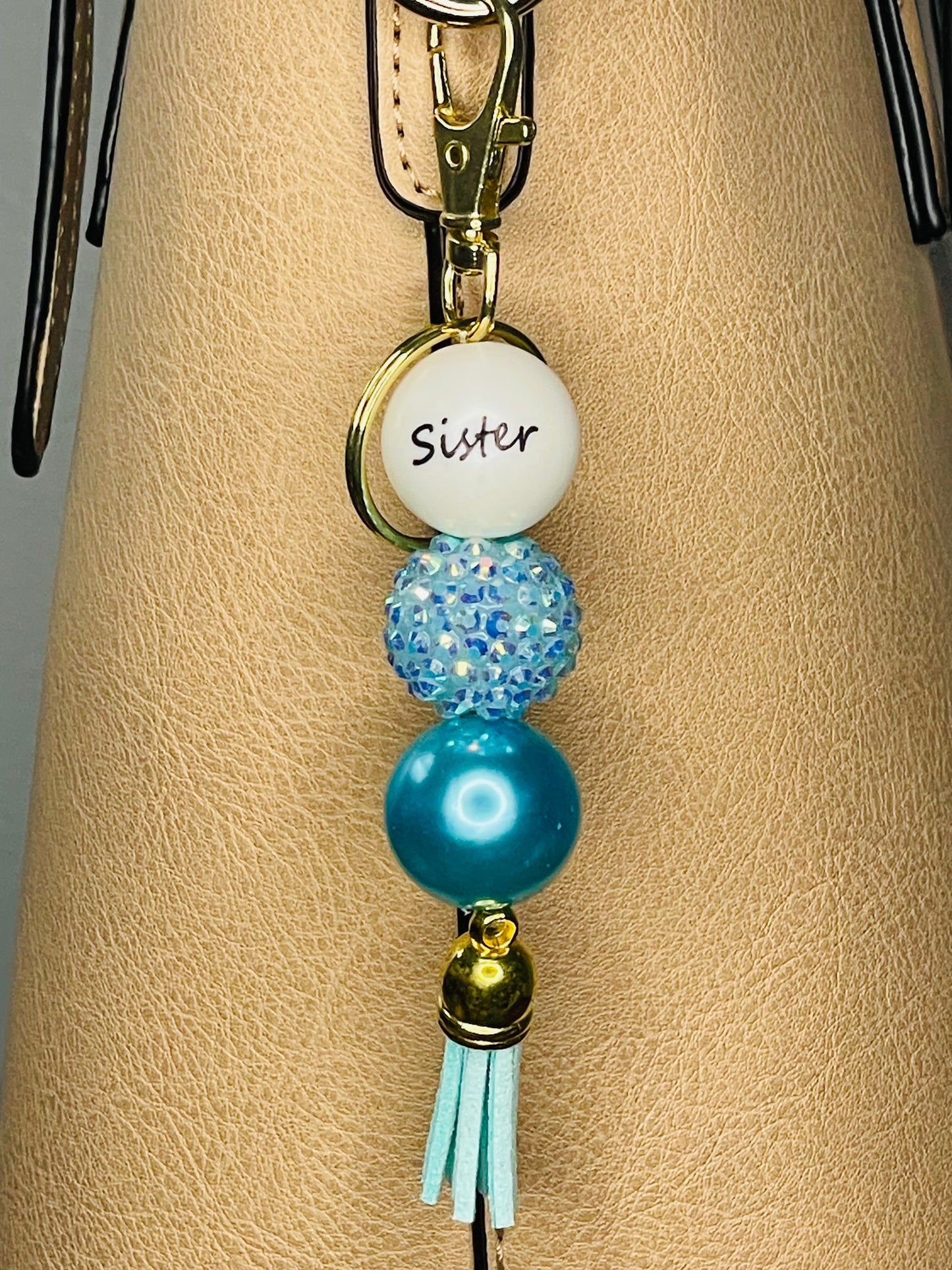 A key ring on a gold ring with 3 beads, the first white with Sister written on the bead, the second is a blue rhinestone bead, the third is an iridescent teals pearl bead. The key chain is finished with a light teal tassel.
