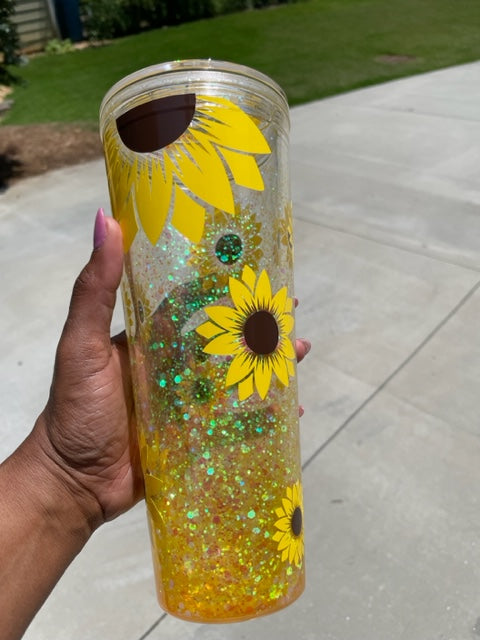 A clear double walled  acrylic cup decorated with sunflowers. The space between the double walls is filled with  yellow glitter and a solution to make the glitter flow and move.