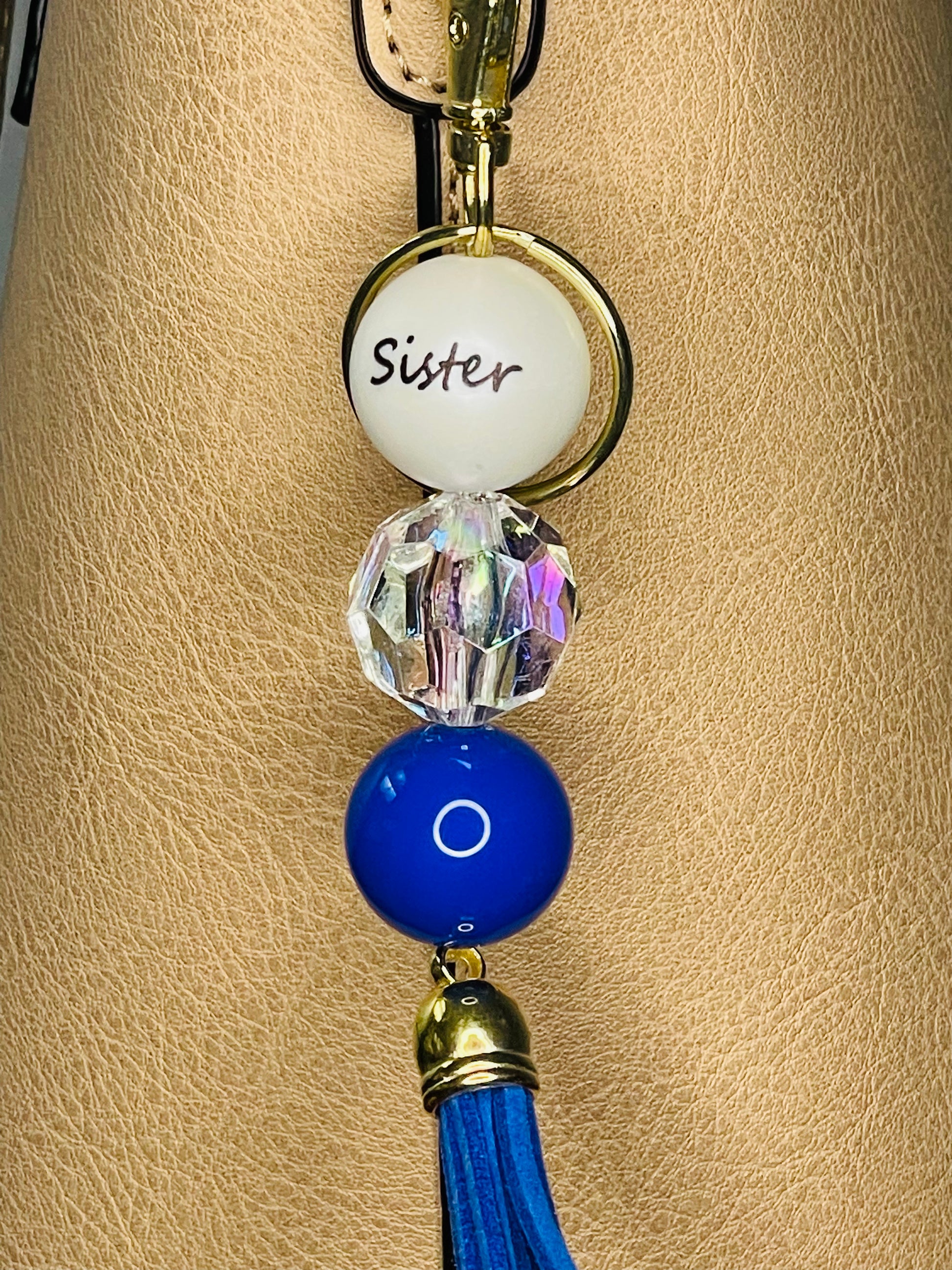 A key ring on a gold ring with 3 beads, the first white with Sister written on the bead, the second is a facetted bead, the third is a royal blue bead. The key chain is finished with a blue tassel.