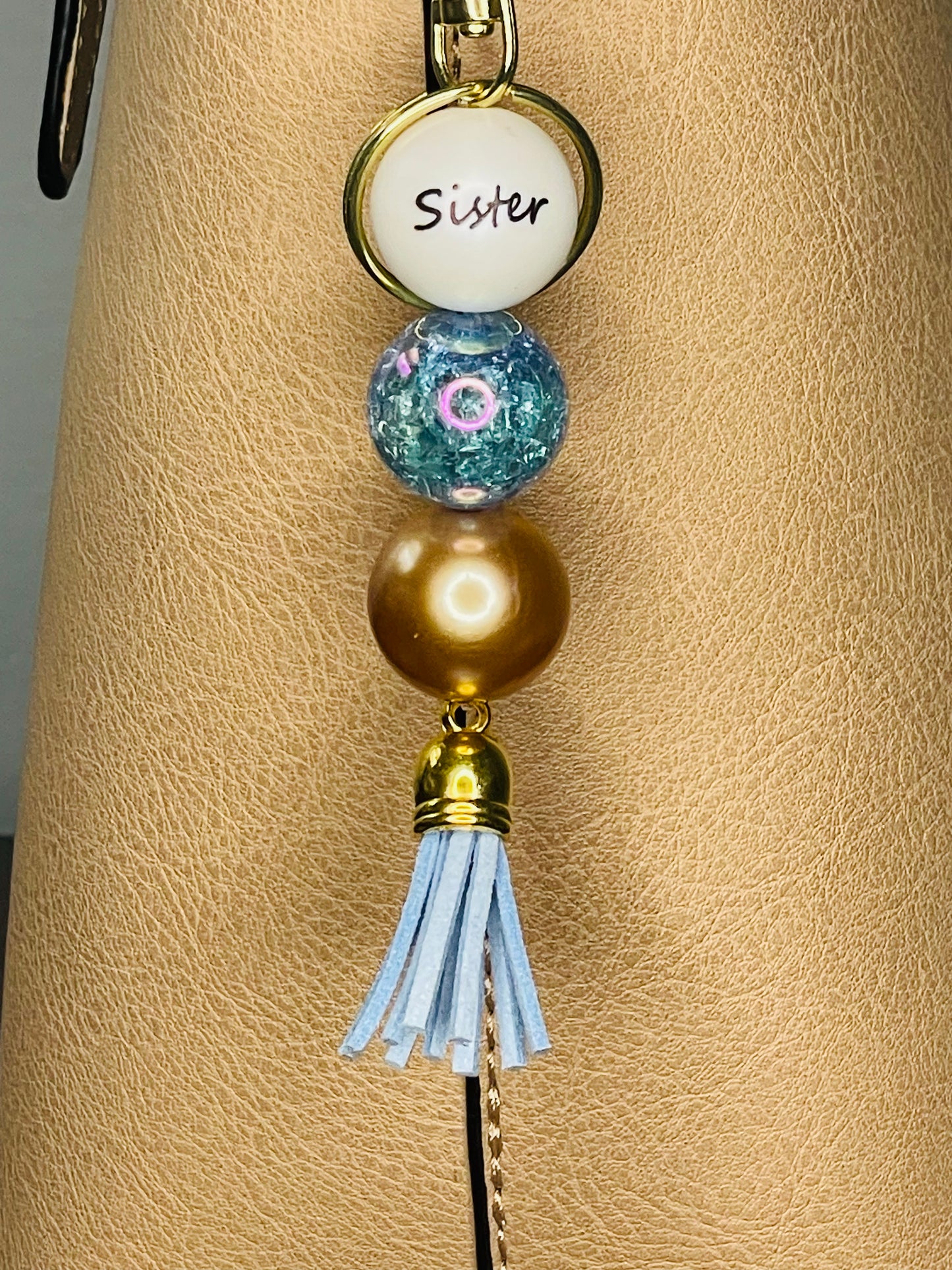 A key ring on a gold ring with 3 beads, the first white with Sister written on the bead, the second is a blue  clear sparkled blue bead, the third is a gold bead. The key chain is finished with a light blue tassel.