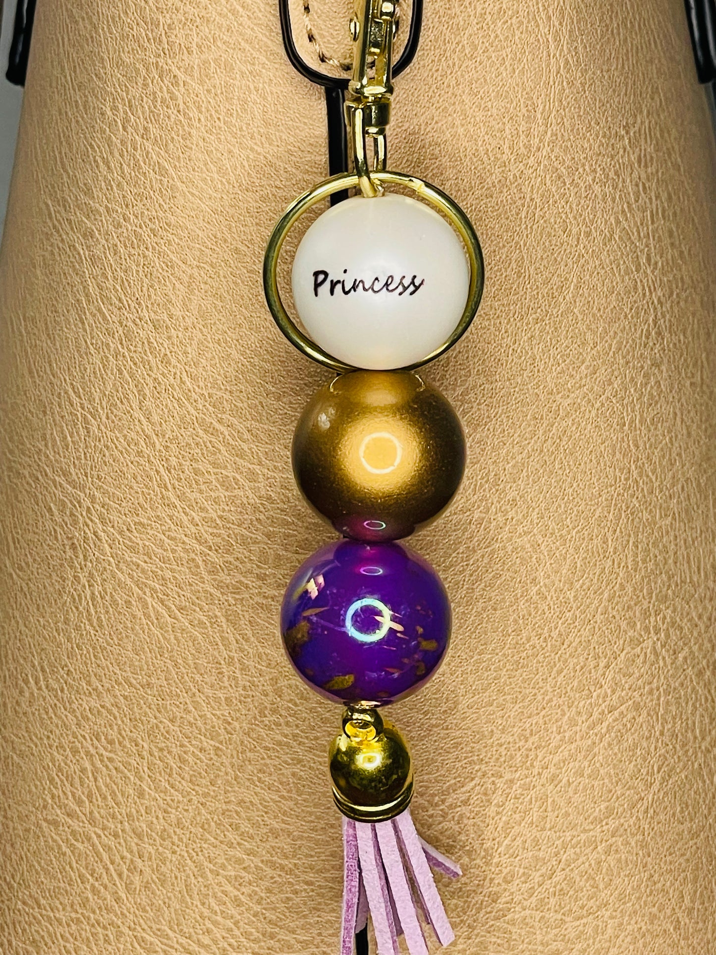 A key ring on a gold ring with 3 beads the first white with Princess written on the bead, is a deep shiny gold , the third a  dark purple with gold fleck swirls. The key chain is finished with a light purple tassel