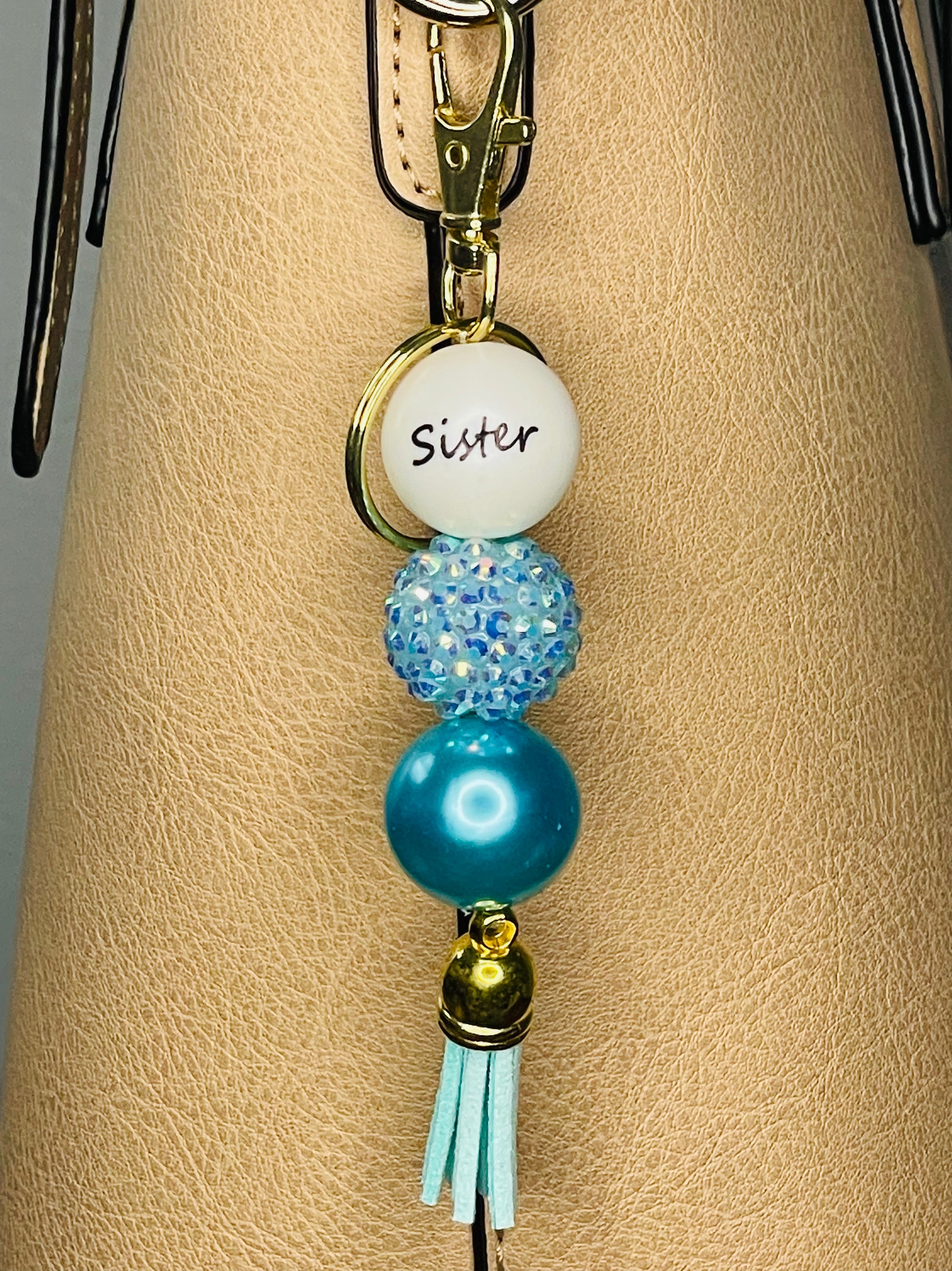 A key ring on a gold ring  with 3 beads, the first white with Sister written on the bead, the second is a blue rhinestone bead, the third is an iridescent teals pearl bead. The key chain is finished with a light teal tassel.