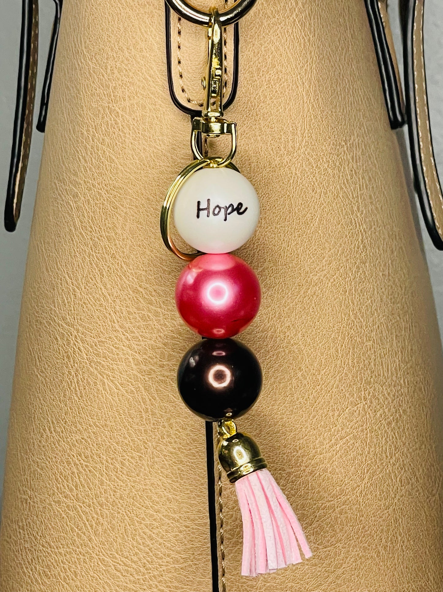 A key ring on a gold ring with 3 beads the first white with hope written on the bead, the second is satin deep pink, the third a deep chocolate shiny brown. The key chain is finished with a pink tassel.