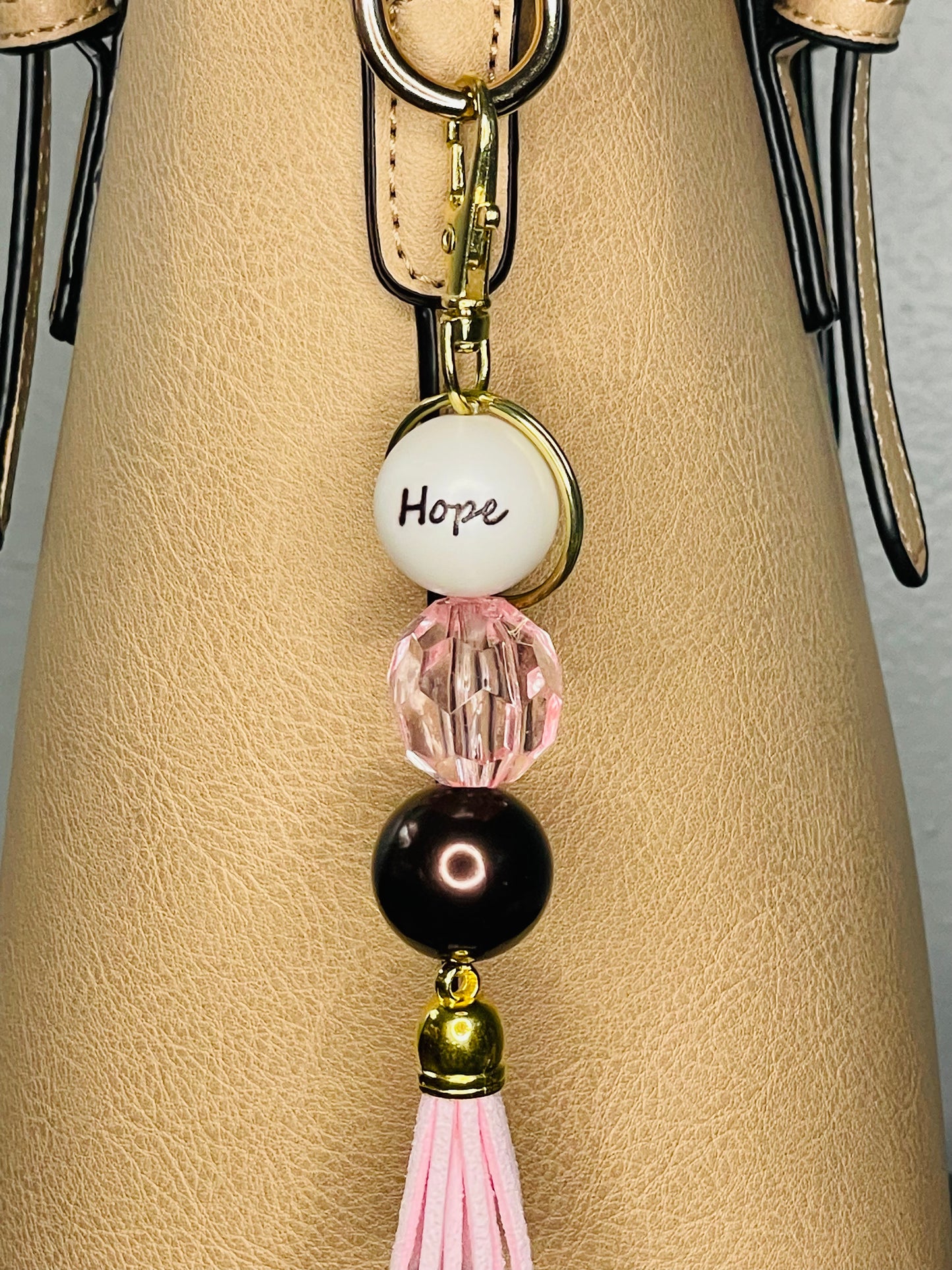 A key ring on a gold ring with 3 beads the first white with hope written on the bead, the second is clear pink facetted, the third a deep chocolate shiny brown.A closer view. The key chain is finished with a pink tassel.